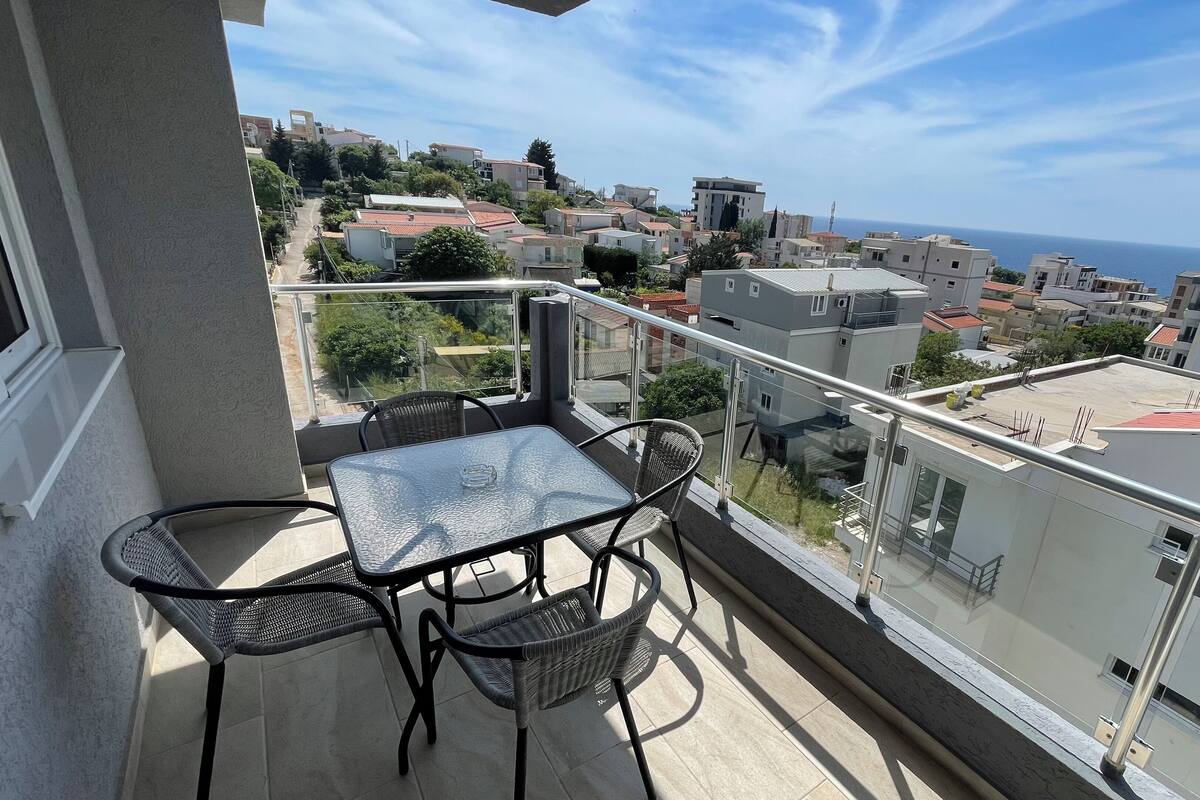 A balcony with a table and chairs, overlooking a coastal town with buildings and the sea.