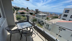Varanda com mesa e cadeiras, com vista para uma cidade costeira com edifícios e o mar.