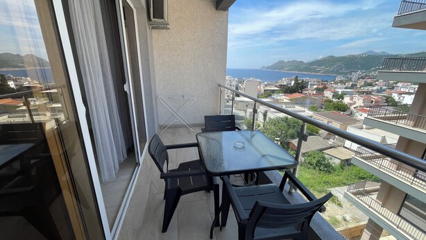 A balcony with a glass table and chairs, overlooking a coastal cityscape with buildings and mountains.