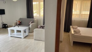 A modern living room with a grey sofa, a white coffee table, and a bed with neatly folded towels. - Elma Hotel (Bar)