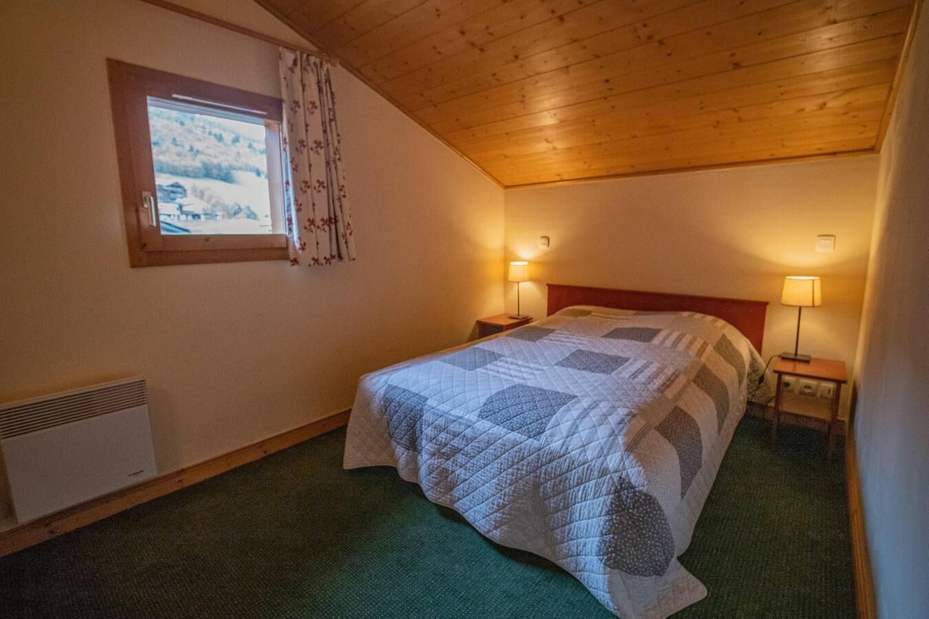 A bedroom with a wooden ceiling, a bed with a quilt, two bedside lamps, a window with curtains, and a radiator.