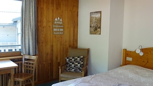 Une pièce aux murs lambrissés, contenant un lit, un bureau et une chaise. Un panneau avec du texte est accroché au mur, ainsi qu’un tableau représentant un cerf.
