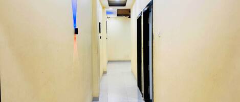 A narrow, well-lit hallway with a tiled floor, leading to a room with a black door and a ceiling fan.