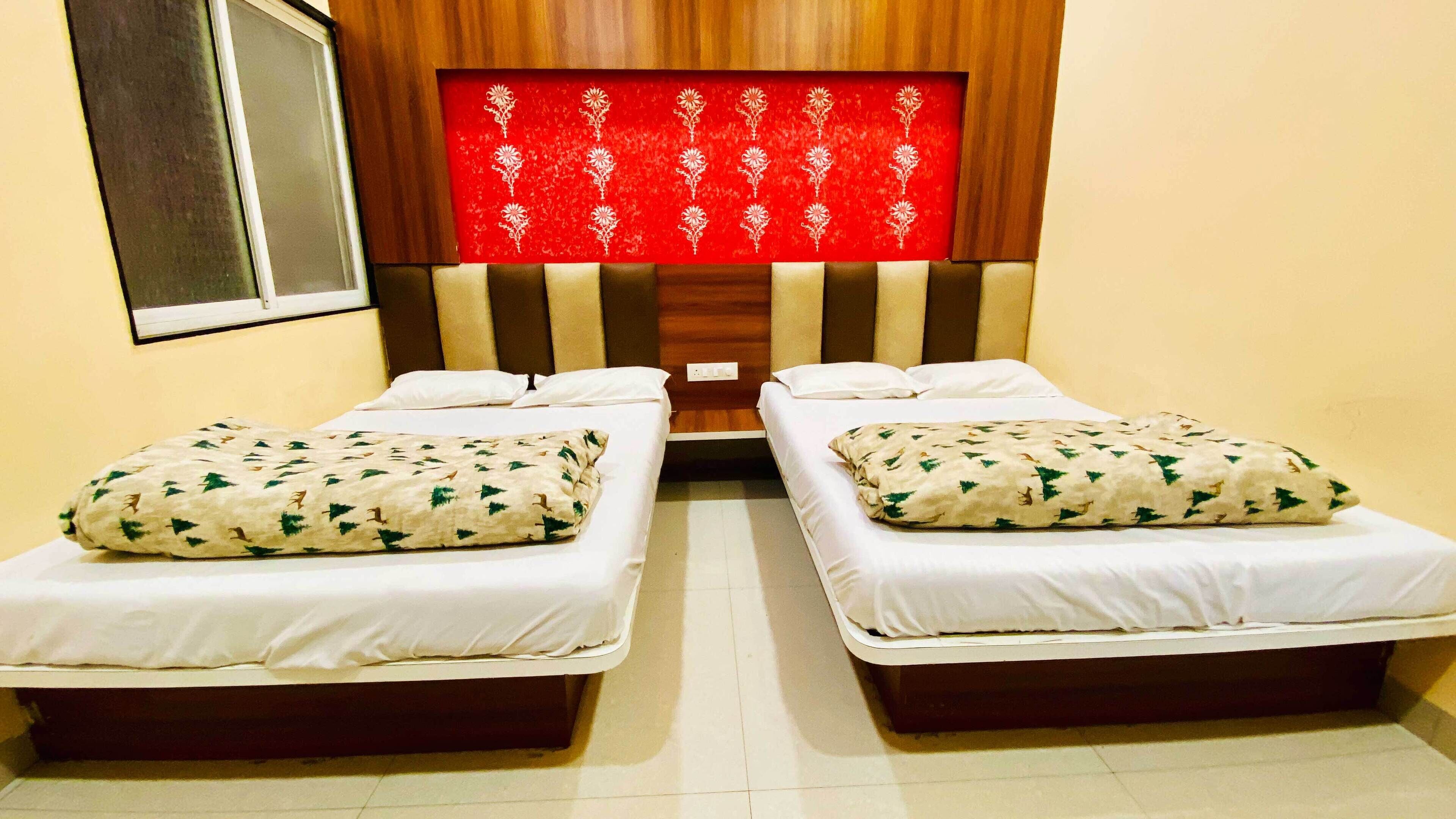A hotel room with two single beds, each with a white mattress and a patterned pillow. The headboard is wooden with a red decorative panel.