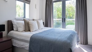 Een slaapkamer met een groot bed, een raam met gordijnen, een nachtkastje en uitzicht op groen.
