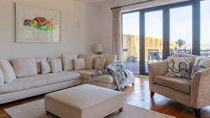 Een moderne woonkamer met een hoekbank, een chaise-lounge en een bijzettafeltje. Er is een groot raam met uitzicht op de omgeving.
