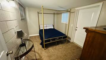 A bedroom with a four-poster bed, a small round table with a lamp, a wooden dresser, and a white door.