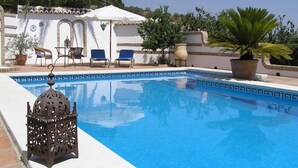 A swimming pool with blue tiles, surrounded by palm trees and potted plants. There are lounge chairs and a decorative metal lantern near the pool.