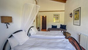 A room with two beds, a wooden ceiling, a wall-mounted lamp, a framed picture, and a door leading to another room.