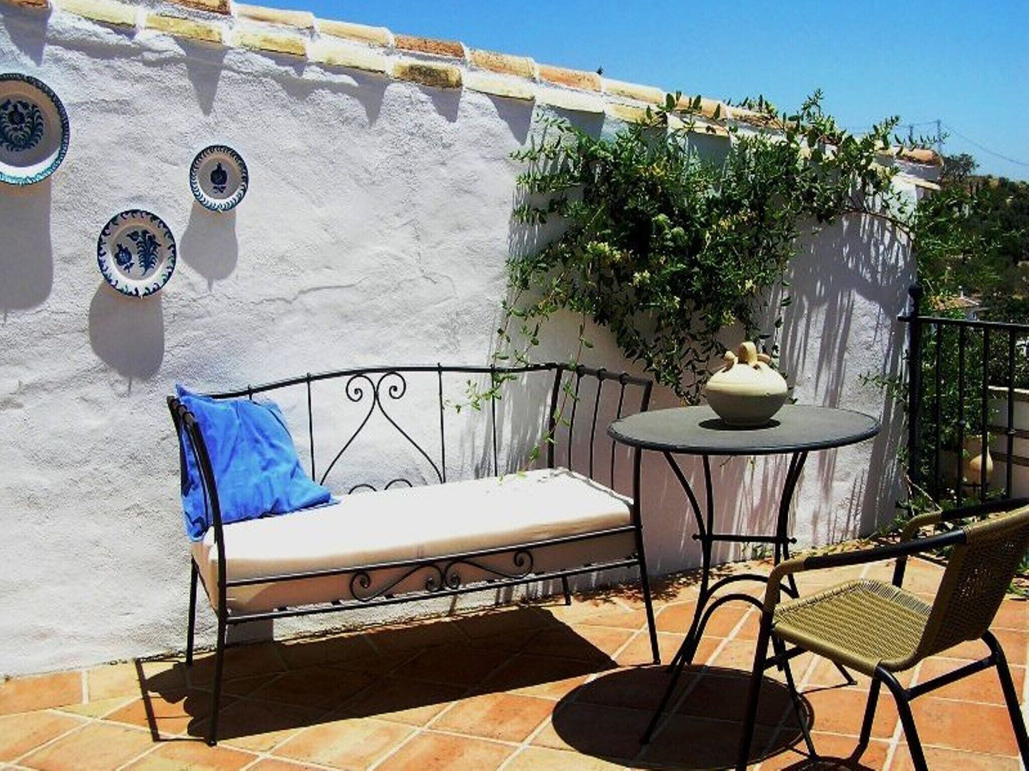 A patio with a white wall, a blue cushion, a small round table with a white bowl, and a wicker chair.