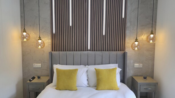 A modern bedroom with a grey upholstered headboard, a large bed with white and yellow pillows, and two bedside tables with lamps.