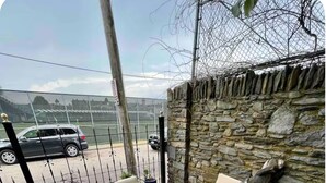 Patio esterno con tavolo rotondo in vetro e sedie in metallo bianco, circondato da un muro in pietra e una recinzione metallica.
