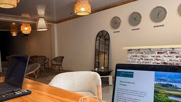 A room with a wooden table, two laptops, a glass of wine, and a laptop displaying a webpage. There are wicker lamps, a mirror, and clocks on the wall.