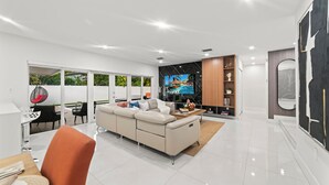A modern living room with a sofa, a coffee table, and a TV. There is a dining area with a table and chairs. The room has a large window and a view of greenery outside. - Family Retreat: Pool, Games & Private Apartment (Miami)
