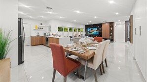 A modern kitchen with white cabinets, a dining area with a wooden table, and a TV displaying a tropical scene. - Family Retreat: Pool, Games & Private Apartment (Miami)