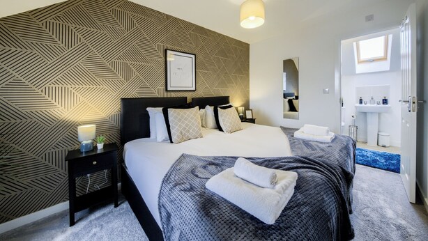 Een moderne slaapkamer met een wand met een geometrisch patroon, een net opgemaakt bed met handdoeken, een nachtkastje met een lamp en een badkamer zichtbaar door een geopende deur.