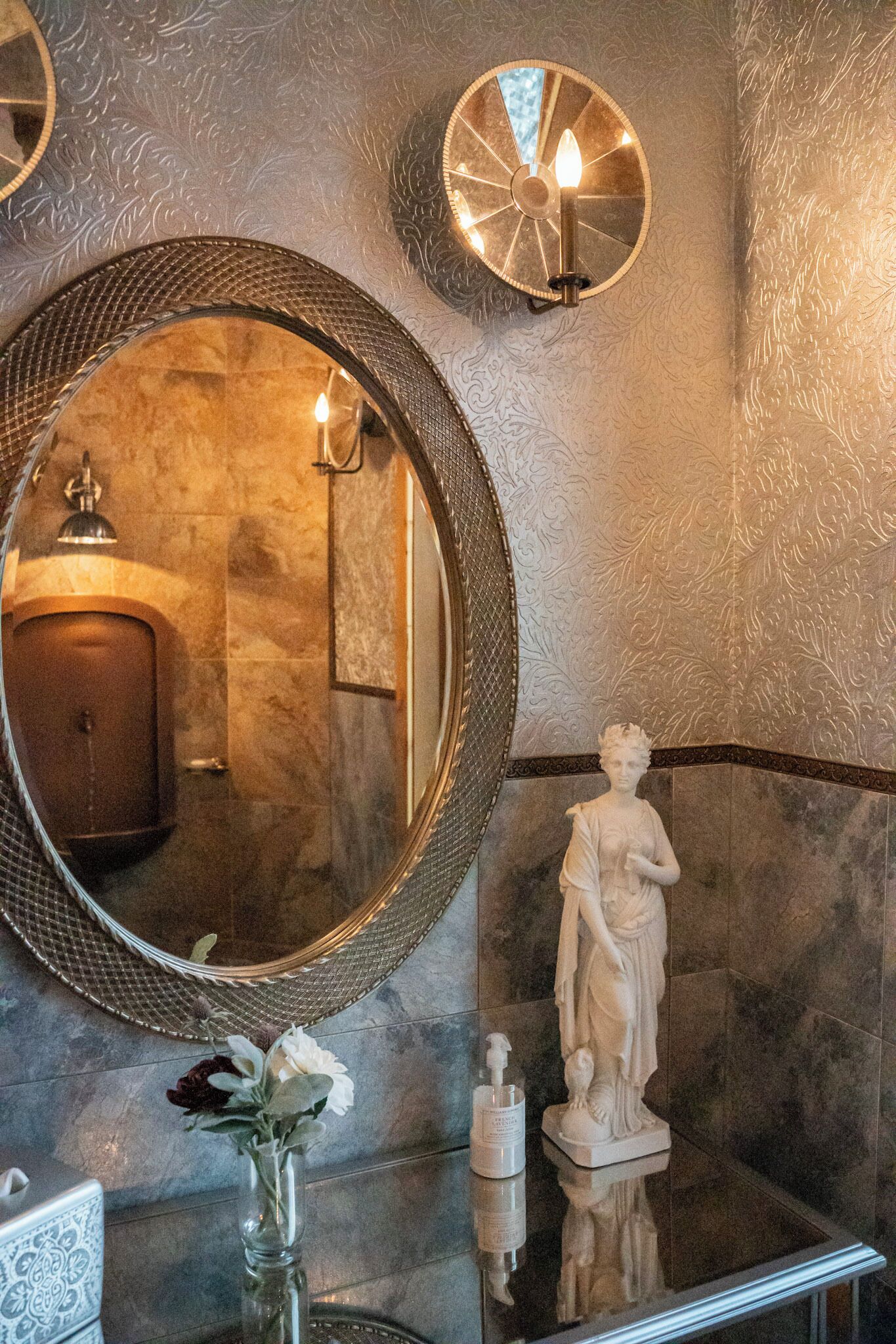 Une salle de bain avec un grand miroir rond, une statue et un vase rempli de fleurs.
