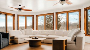 A modern living room with a sectional sofa, a coffee table, and a ceiling fan. Large windows provide a view of the outdoors.