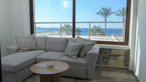 A modern living room with a grey sectional sofa, a round wooden coffee table, and a large window offering a view of the sea and palm trees.