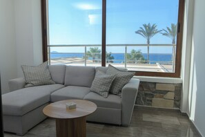 A modern living room with a grey sectional sofa, a round wooden coffee table, and a large window offering a view of the sea and palm trees. - Envy Seaside Suites (Heraklion)