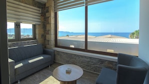 A room with a stone wall, a grey sofa, a round table, and a large window with a view of the sea and buildings. - Envy Seaside Suites (Heraklion)