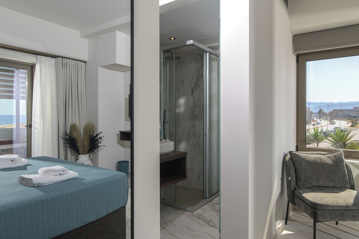 A modern hotel room with a bed, a shower area, a balcony with a view, and a seating area.