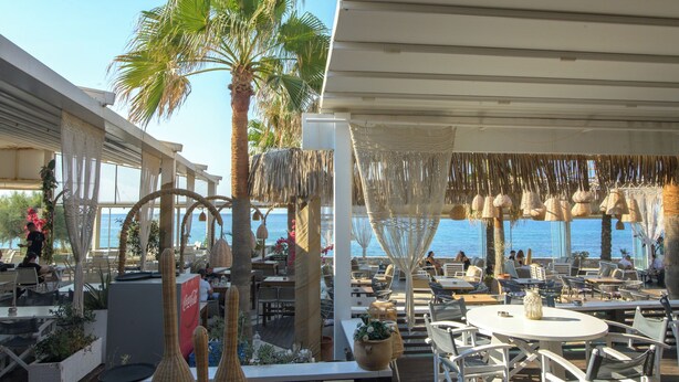 An outdoor dining area with tables and chairs, palm trees, and a view of the sea. - Envy Seaside Suites (Heraklion)