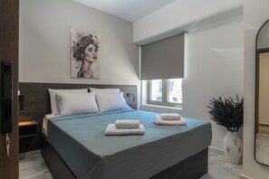 A modern bedroom with a large bed, a bedside table, a wall-mounted TV, a window with a blind, and a decorative vase. - Envy Seaside Suites (Heraklion)
