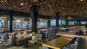 A modern restaurant with wooden ceiling panels, multiple seating areas including booths and tables, and large windows providing views of the outside. - Envy Seaside Suites (Heraklion)