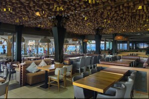 A modern restaurant with wooden ceiling panels, multiple seating areas including booths and tables, and large windows providing views of the outside. - Envy Seaside Suites (Heraklion)