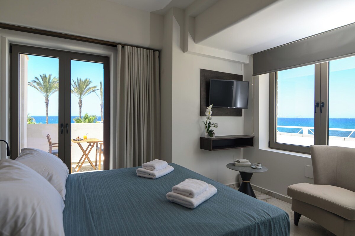 A hotel room with a bed, a TV, a small table, a chair, and a balcony with a view of the sea.