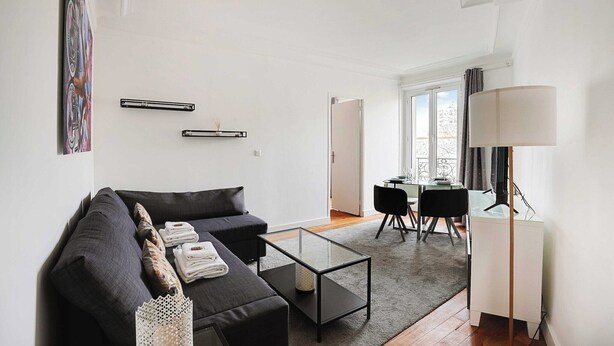 A modern living room with a dark sofa, a glass coffee table, and a dining area with a table and chairs. - Chic apartment - 1BR/4P - Next to Paris (Saint-Ouen-sur-Seine)