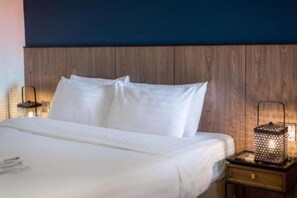 Una cama bien hecha con sábanas y almohadas blancas, cabecera de madera y mesita de noche con una linterna encendida.