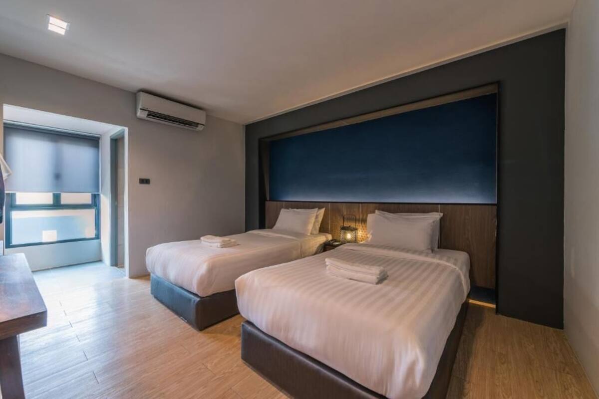A modern hotel room with two beds, a wooden floor, a large window, and a dark accent wall.