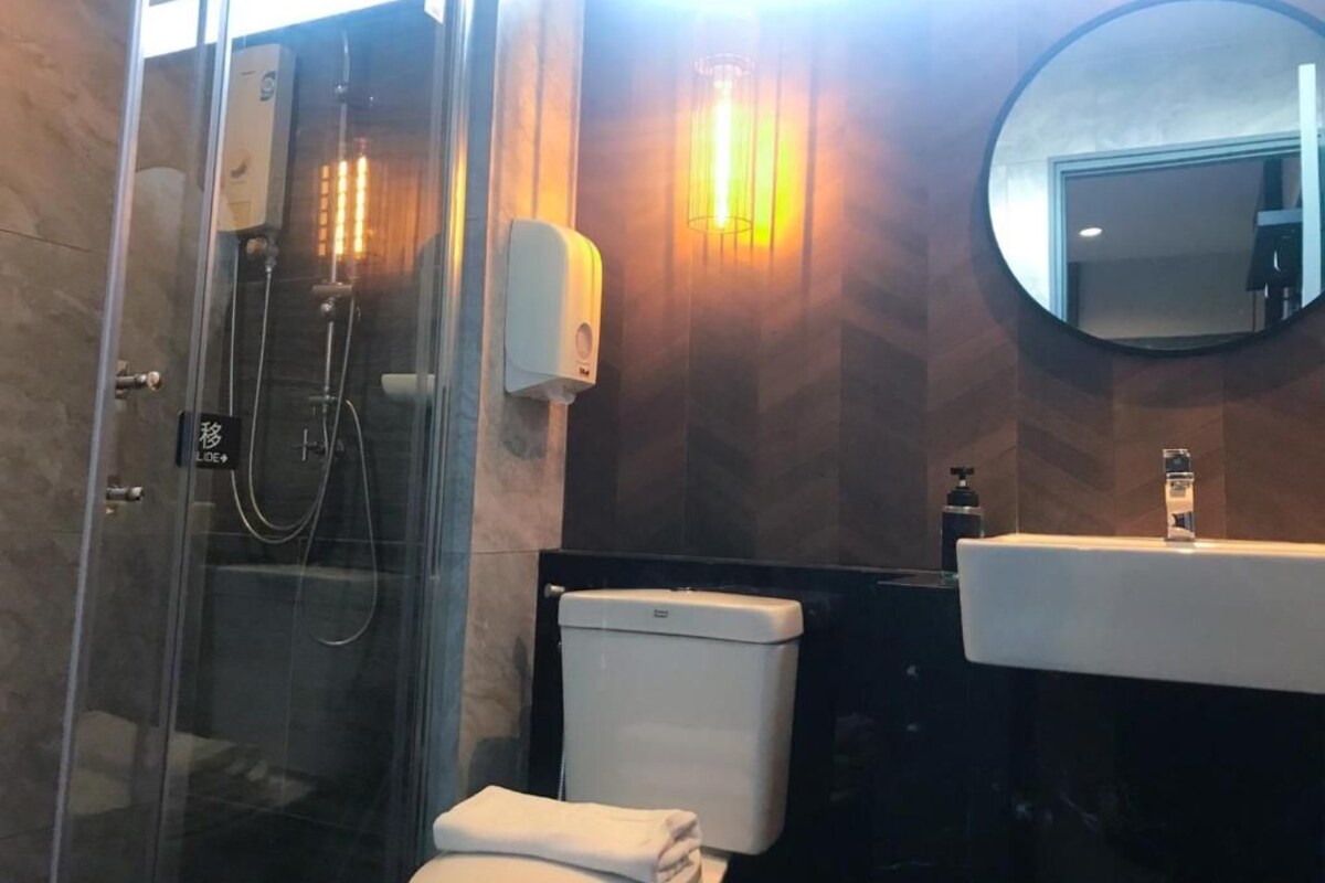 Un baño moderno con ducha de vidrio, inodoro blanco, espejo redondo y dispensador de jabón en la pared.