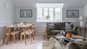 A cozy living room with a dining area, featuring a bay window, a sofa with patterned cushions, and a coffee table with oranges.