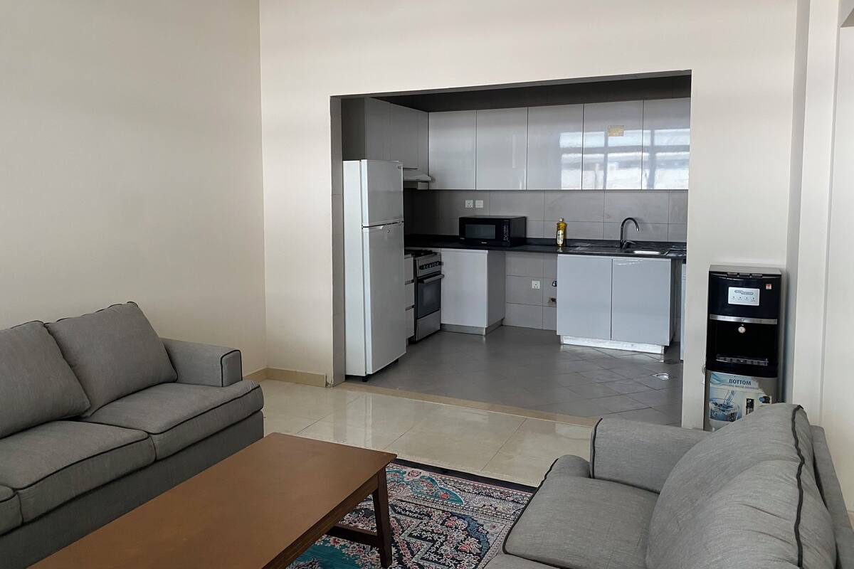 Una cocina moderna con armarios blancos, un refrigerador, microondas y dispensador de agua. En la sala de estar, hay un sofá gris y una mesa de centro.