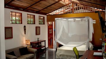 Quarto com cama, sofá, escrivaninha de madeira, computador e janela com vista para o verde.