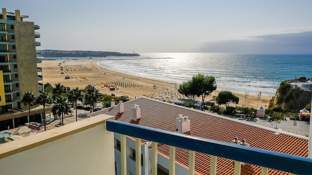 A beach view from a balcony with a tiled roof, palm trees, and a building with balconies. - Apartament Sea view Concorde (Portimão)