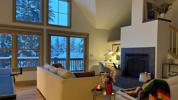 A cozy living room with a sofa, a small table with a lamp, a fireplace, and large windows overlooking a snowy deck. - Spacious 3BR Retreat with Mt. Views, Walk to the lake. 30 days stay! (Whitefish)