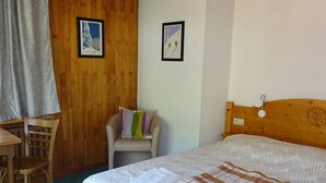 A room with a bed, a chair, a desk, and two framed pictures on the wall. - Spacious 2-Room Apt in Les Gets Center - Sleeps 4, WiFi, Linens Included, Near Ski Lifts (Les Gets)