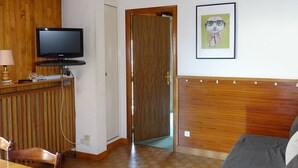A room with a television on a wooden stand, a framed picture on the wall, a wooden door, and a tiled floor. - Spacious 2-Room Apt in Les Gets Center - Sleeps 4, WiFi, Linens Included, Near Ski Lifts (Les Gets)