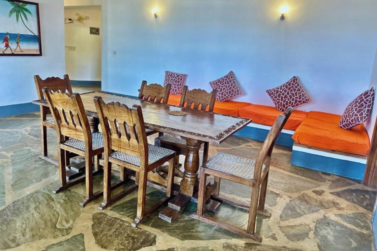 Un comedor con una mesa y sillas de madera, un sofá con cojines estampados y una pared decorada con un cuadro.