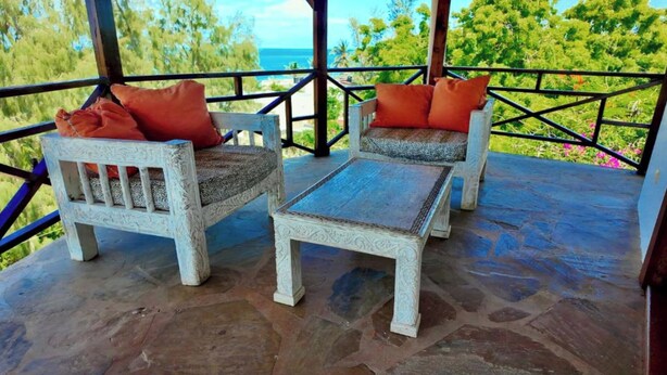 A covered outdoor seating area with two white wooden chairs and a small table, featuring orange cushions. - Silver Rock Hotel (Malindi)