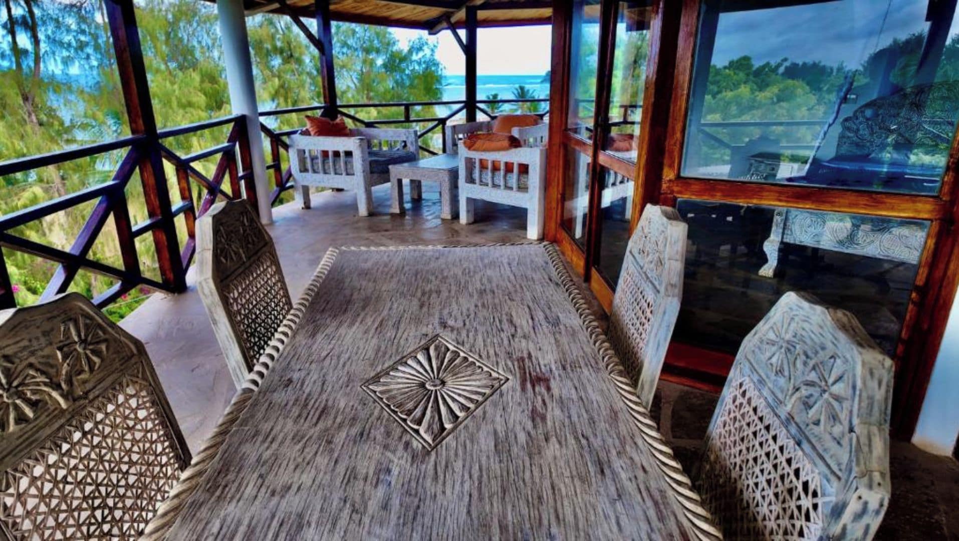 Un espacio al aire libre cubierto con mesa y sillas de madera, rodeado por una barandilla y vista al océano.