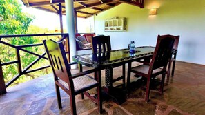 A covered outdoor dining area with a table and chairs, a water bottle on the table, and a view of greenery outside.