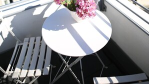 A balcony with a white table and chairs, a flower vase with pink flowers, and a view of a town with red-roofed buildings.