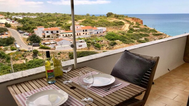 A balcony table with plates, wine glasses, and a bottle, overlooking a coastal town with buildings and a clear sky.