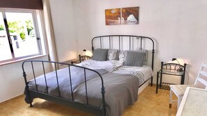 A bedroom with a metal bed frame, a bedside table with a lamp, a chair, and a window with curtains.
