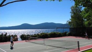 An outdoor tennis court with a net, surrounded by a fence and greenery, overlooking a body of water and mountains. - Lakefront Condo in Kings Beach- #12 (Kings Beach)